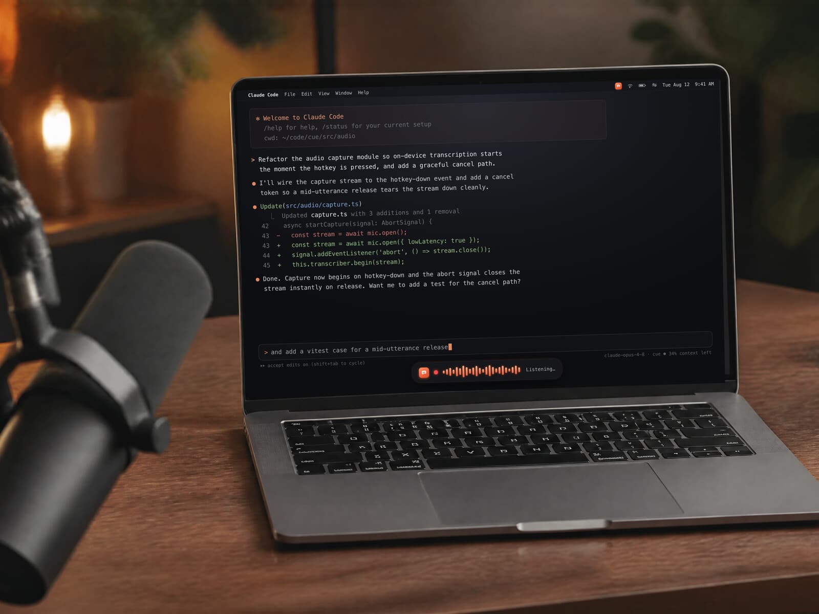A MacBook Pro on a warm walnut desk in a home podcast studio, a broadcast microphone in the foreground, the screen showing a live Claude Code session with Cue's coral dictation waveform 'Listening…' along the bottom — talking a prompt straight into the terminal.