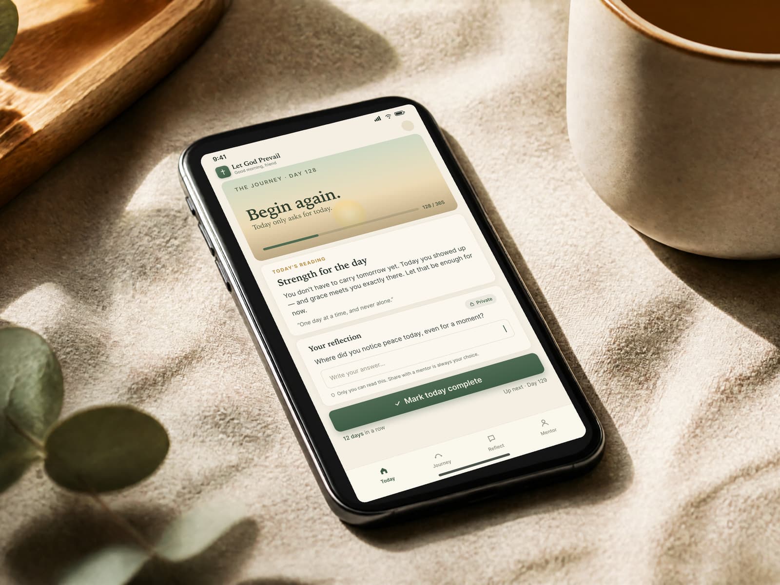 A phone resting on warm linen in soft morning light, beside eucalyptus and a cup of tea — the Let God Prevail companion app open to the day's guided reading, a year-long journey progress bar, and a private, locked reflection space.